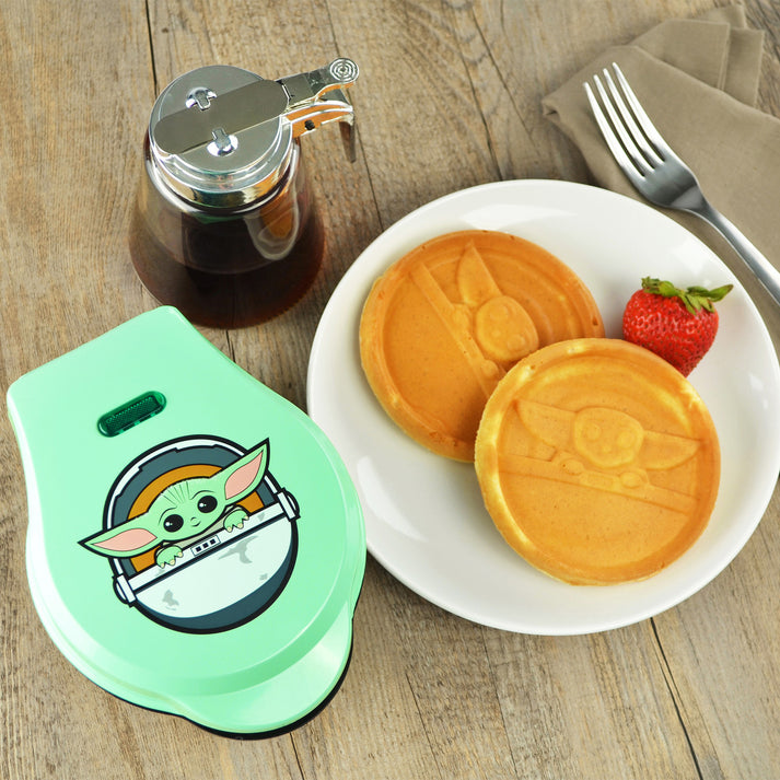 A green waffle maker next to a plate of two waffles and a syrup container